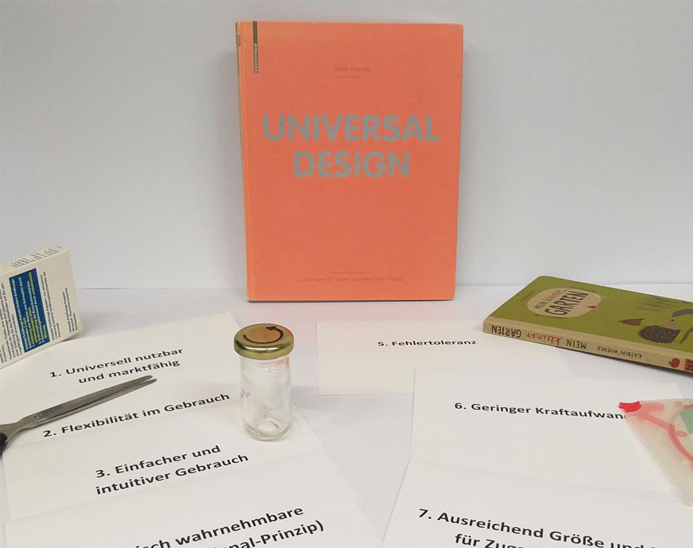 Ein Buch über Universal Design steht an der Wand gelehnt und davor liegen diverse Arbeitsmaterialien sowie Karteikarten, mit den 7 Punkten des universellen Designs.