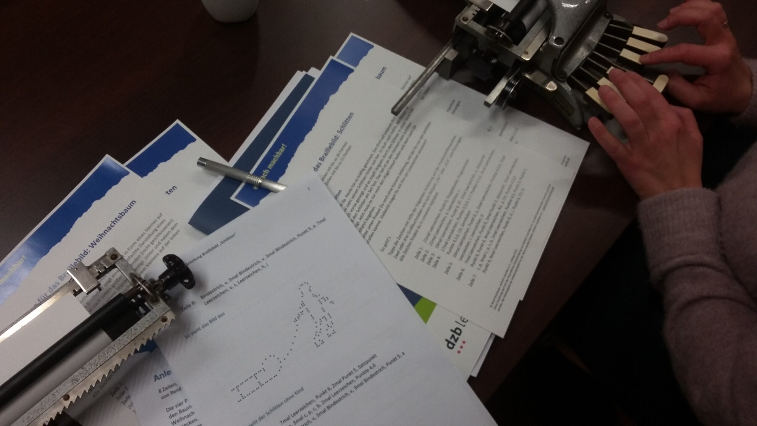 Workshopunterlagen und Arbeitsmaterialien liegen auf dem Tisch neben einer Punktschriftmaschine.