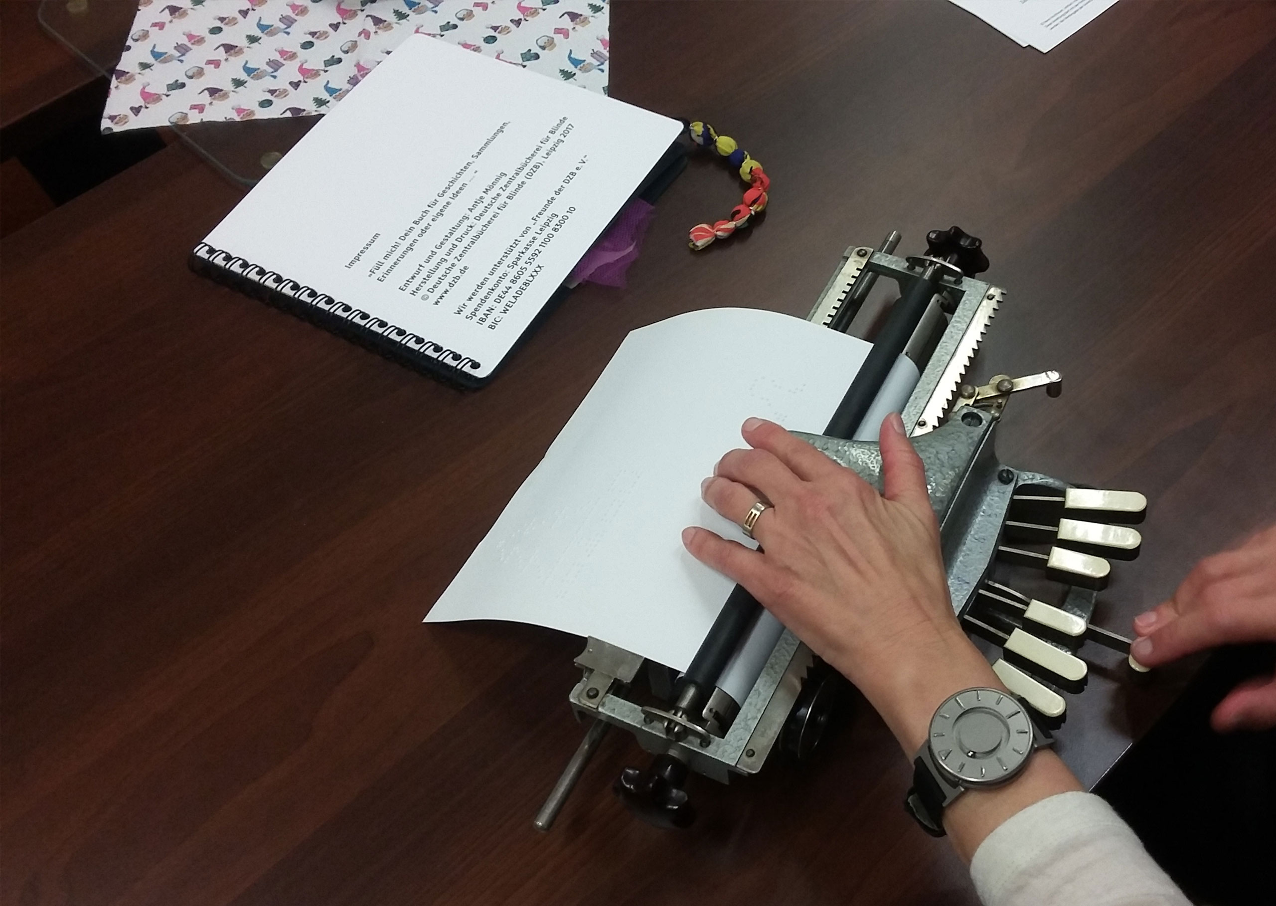 Eine Person schreibt mit einer Punktschriftmaschine. Die Hände sind zu sehen. Eine auf den Tasten, eine auf dem Blatt.