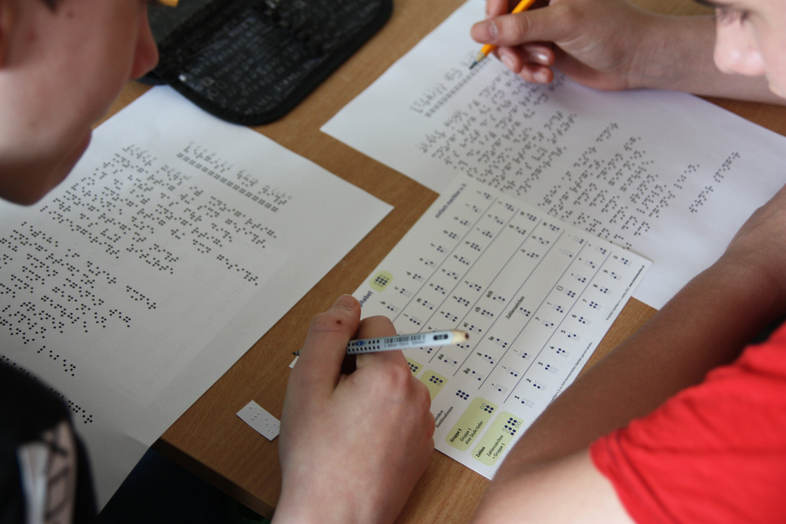 Zwei Jugendliche sitzen an einer Schulbank mit Zettel, Stift und Braille-Alphabet, um das Braille-Gedicht in Schwarzschrift zu übertragen.